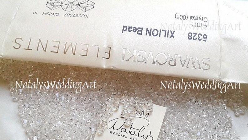 May include: A collection of clear, faceted 4 mm Swarovski XILION beads. The beads are displayed alongside their original white packaging, which features the text SWAROVSKI ELEMENTS, 5328 XILION Bead, 4 mm Crystal (001), and 103557987.