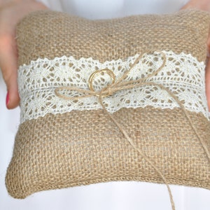 May include: A burlap ring pillow with a white lace trim and a gold ring tied in the center with twine.