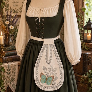 Puede incluir: Un vestido verde oscuro con una blusa color crema de manga larga y escote cuadrado. El corpiño tiene un frente con cordones y un delantal color crema está sujeto a la cintura, decorado con bordados florales negros y una mariposa azul.