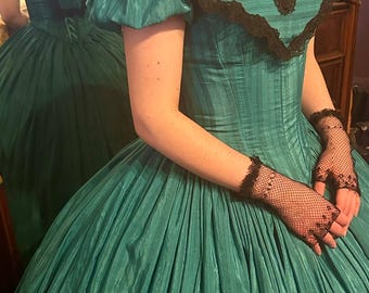 May include: A teal green ball gown with a black lace trim around the neckline and sleeves. The gown has a fitted bodice and a full skirt. The model is wearing black lace gloves.