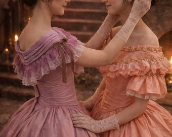 Puede incluir: Dos mujeres con vestidos de época, uno rosa empolvado y otro melocotón, están juntas. Los vestidos tienen escotes descubiertos, detalles de encaje y faldas amplias. Ambas llevan adornos florales en el pelo y guantes de encaje, creando una estética romántica e histórica.