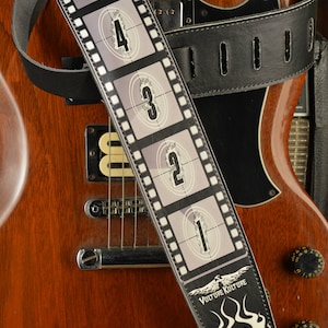 May include: Black leather guitar strap with a white film strip design featuring numbers 5 through 1. The strap has a white flame design with the text "Vulture Culture" at the bottom.