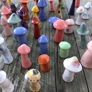 May include: A collection of colourful ceramic mushroom figurines. The mushrooms are painted in various shades of blue, green, pink, red, and yellow. Some of the mushrooms have white speckles or dots.