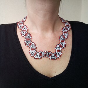 Puede incluir: Un collar de cuentas con un patrón geométrico en negro, blanco y rojo. El collar lo lleva una persona con una camisa negra.