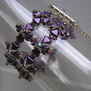 Puede incluir: Un brazalete de cuentas moradas y plateadas con cierre de broche. El brazalete está hecho de pequeñas cuentas triangulares que están dispuestas en un patrón de flores.
