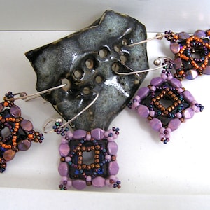 Puede incluir: Tres pares de pendientes de cuentas moradas, marrones y naranjas con ganchos de plata. Los pendientes tienen forma de cuadrado con un cuadrado más pequeño en el centro.