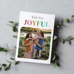 May include: A white card with a family photo and the word "JOYFUL" in rainbow colors. The family photo shows a smiling family of four. The card is decorated with green leaves.