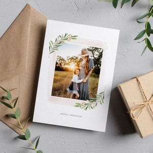 May include: A Christmas card featuring a watercolour-style illustration of greenery and berries framing a photograph of a mother and daughter. The photo depicts the mother and daughter smiling and embracing in a field. The card reads "Merry Christmas".