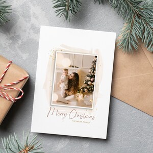 Puede incluir: Una tarjeta de Navidad con una foto de una madre y un ni&ntilde;o frente a un &aacute;rbol de Navidad decorado. La tarjeta tiene un fondo blanco con purpurina dorada y el texto "Feliz Navidad La familia Mark".