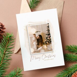 Puede incluir: Una tarjeta de Navidad con una foto de una madre y un ni&ntilde;o frente a un &aacute;rbol de Navidad decorado. La tarjeta tiene un fondo blanco con un dise&ntilde;o de acuarela y detalles de purpurina dorada. El texto "Feliz Navidad" y "La familia Mark" est&aacute; impreso en marr&oacute;n en la tarjeta.
