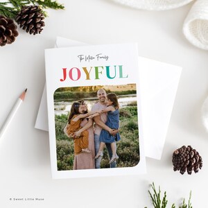 May include: A white greeting card with the word "JOYFUL" in rainbow colors and the family name in black script. The card features a family photo inside a rounded rectangle. A white envelope is behind the card.