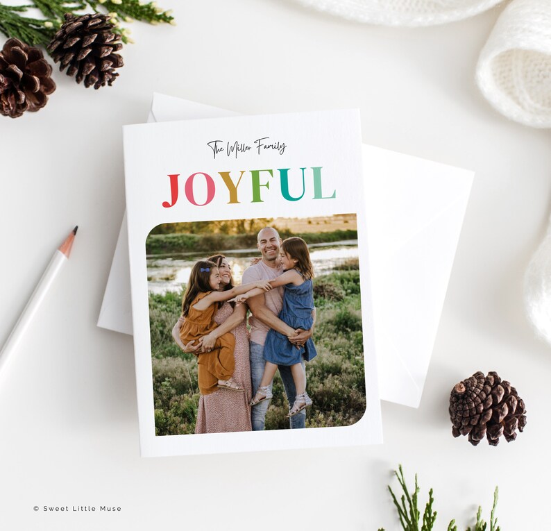 May include: A white greeting card with the word "JOYFUL" in rainbow colors and the family name in black script. The card features a family photo inside a rounded rectangle. A white envelope is behind the card.