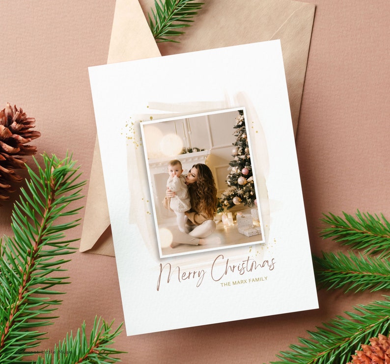 Puede incluir: Una tarjeta de Navidad con una foto de una madre y un ni&ntilde;o frente a un &aacute;rbol de Navidad decorado. La tarjeta tiene un fondo blanco con un dise&ntilde;o de acuarela y detalles de purpurina dorada. El texto "Feliz Navidad" y "La familia Mark" est&aacute; impreso en marr&oacute;n en la tarjeta.