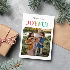 May include: A holiday card with the word "JOYFUL" in rainbow colors, featuring a family photo. A wrapped gift with red and white twine and evergreen branches are in the background. A brown envelope is also visible.