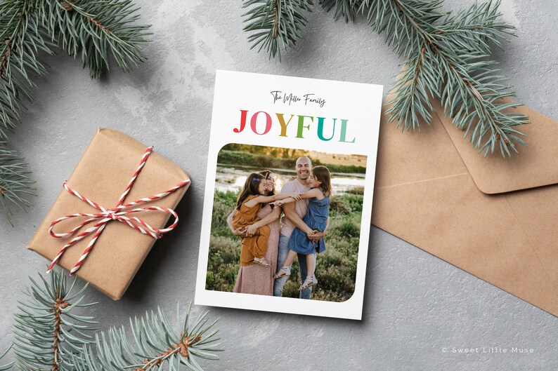 May include: A holiday card with the word "JOYFUL" in rainbow colors, featuring a family photo. A wrapped gift with red and white twine and evergreen branches are in the background. A brown envelope is also visible.
