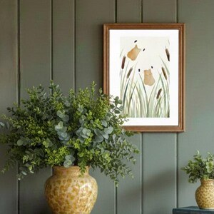 May include: A framed watercolor print of cattails and two brown shapes, likely animals, in a field of green grass. The print is in a wooden frame. A yellow vase with a textured pattern holds green foliage. A smaller vase with foliage is also present.