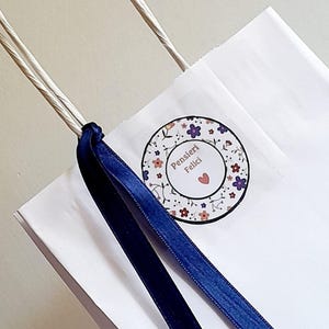 May include: A white paper gift bag with twisted paper handles, featuring a circular sticker with a floral pattern and the text Pensieri Felici with a small red heart. A dark blue satin ribbon is tied around one of the handles of the bag.