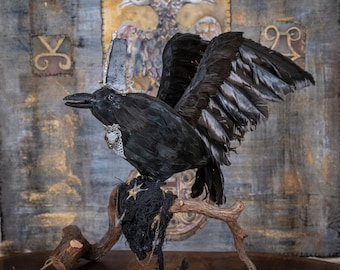 May include: A black feathered raven figurine perched on a wooden branch, wearing a tall silver crown and a silver chain necklace. The bird has its wings partially spread, and a dark, textured fabric nest is attached to the branch beneath it.