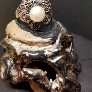May include: A silver ring with a large white pearl set in a delicate floral design. The ring is made of marcasite and has a vintage look.