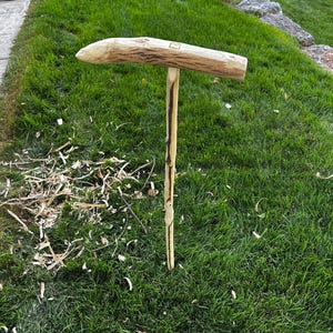 May include: A handcrafted wooden tool with a T-shaped design. The horizontal piece is a light brown color with a textured surface, while the vertical piece is a lighter shade of brown. The tool is set in a bed of green grass.