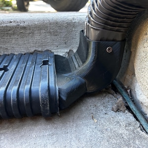 May include: A black 3D-printed gutter downspout extension adapter connecting a corrugated plastic downspout to a rectangular drainage pipe. The adapter is secured with a small metal screw and features a ribbed texture on the drainage pipe.