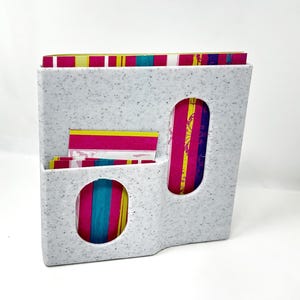 May include: A speckled gray desk organizer with two oval cutouts and a small pocket. The organizer holds colorful striped stationery with pink, yellow, and teal accents. The design is modern and functional.