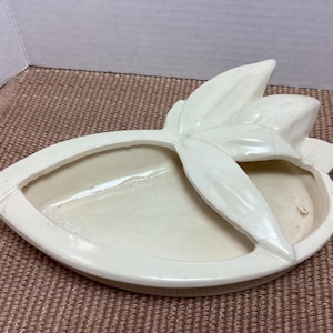 May include: Off-white ceramic decorative dish with a leaf-like design. The dish has a central oval section and a raised leaf-shaped element. The piece is set on a textured, brown surface.