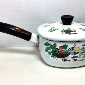 May include: A white enameled metal saucepan with a black handle and a matching lid. The exterior features colorful illustrations of vegetables, herbs, and kitchenware, including a tomato, green pepper, and a basket, with small text labels.