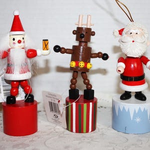 May include: Three wooden Christmas ornaments: Santa Claus, a reindeer, and another Santa Claus. The Santa ornaments are red and white, while the reindeer is brown. Each ornament stands on a cylindrical base, with the Santa ornaments holding small objects.