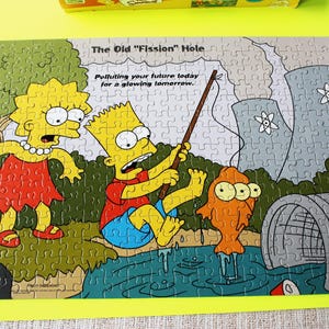 May include: A completed jigsaw puzzle featuring characters from The Simpsons. Bart is fishing a mutated orange fish, with Lisa looking on. The text reads "The Old 'Fission' Hole" and "Polluting your future today for a glowing tomorrow."