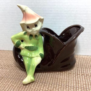May include: A vintage ceramic planter featuring a seated elf figure in green and white attire, with a pink hat. The elf is positioned next to a brown, boot-shaped planter, creating a whimsical decorative piece.
