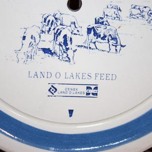 May include: A white ceramic plate with a blue border and a blue and white illustration of cows. The plate has the text "Land O'Lakes Feed" and the logo "CENEX Land O'Lakes" in blue.
