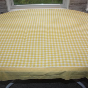 May include: A round tablecloth with a yellow and white gingham pattern. The tablecloth is draped over a table, with a yellow border along the edge. The fabric has a classic, country-style design, suitable for kitchen or dining decor.