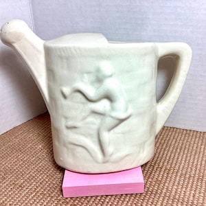 May include: Off-white ceramic watering can with a curved spout and handle. The body features a relief sculpture of figures. The watering can is placed on a pink rectangular block.