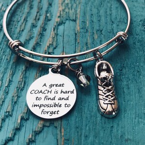 May include: A silver bangle bracelet with a charm that reads "A great COACH is hard to find and impossible to forget". The charm is attached to a silver shoe charm and a clear crystal charm.