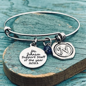 May include: A silver bangle bracelet with a charm that says "Ms. Johnson Support Staff of the year 2021". The bracelet also has a charm with the letter "N" and a small blue crystal.