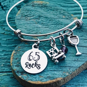May include: Silver charm bracelet with a charm that says "65 Rocks". The bracelet also has a wine glass charm, a chair charm, and a purple gemstone charm.