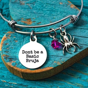 May include: A silver-colored bangle bracelet with three charms: a round pendant with the text "Don't be a Basic Bruja", a small purple crystal, and a spider charm. The bracelet is displayed on a heart-shaped wooden block with a distressed teal finish.