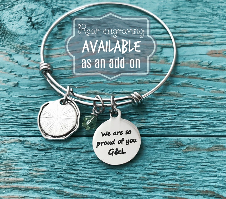Pu&ograve; includere: Braccialetto in argento con un ciondolo rotondo che dice "We are so proud of you G&L". Il braccialetto ha anche un ciondolo in argento con un design a raggiera e un ciondolo di cristallo verde. Il braccialetto &egrave; su una superficie di legno.