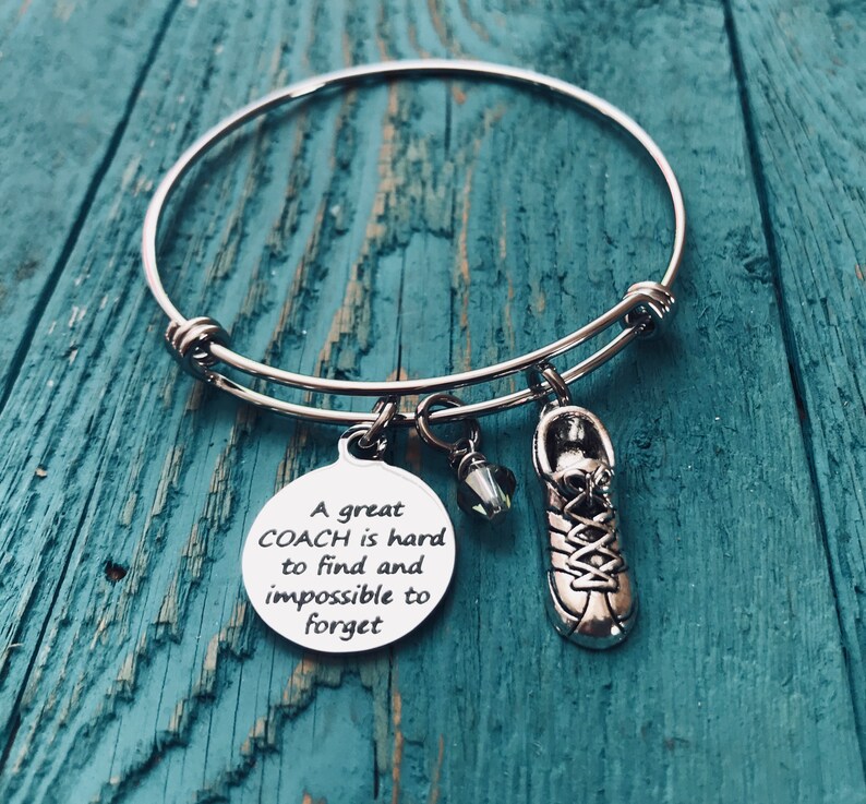 May include: A silver bangle bracelet with a charm that reads "A great COACH is hard to find and impossible to forget". The charm is attached to a silver shoe charm and a clear crystal charm.