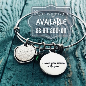 K&ouml;nnte beinhalten: Silbernes Armreif-Armband mit einem Anh&auml;nger, auf dem "Ich liebe dich Mama - Bryan" steht. Das Armband hat au&szlig;erdem einen kleinen gr&uuml;nen Anh&auml;nger und einen runden silbernen Anh&auml;nger mit einem strukturierten Design. Der Text "Gravur auf der R&uuml;ckseite VERF&Uuml;GBAR als Zusatz" wird in einem wei&szlig;en Kasten angezeigt.