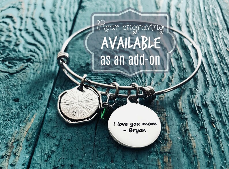 K&ouml;nnte beinhalten: Silbernes Armreif-Armband mit einem Anh&auml;nger, auf dem "Ich liebe dich Mama - Bryan" steht. Das Armband hat au&szlig;erdem einen kleinen gr&uuml;nen Anh&auml;nger und einen runden silbernen Anh&auml;nger mit einem strukturierten Design. Der Text "Gravur auf der R&uuml;ckseite VERF&Uuml;GBAR als Zusatz" wird in einem wei&szlig;en Kasten angezeigt.