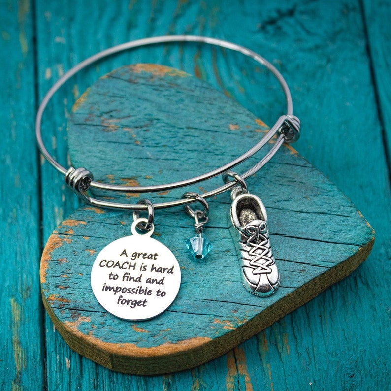 May include: A silver bangle bracelet with a charm that reads "A great COACH is hard to find and impossible to forget". The charm is attached to a small silver shoe charm and a blue crystal charm.