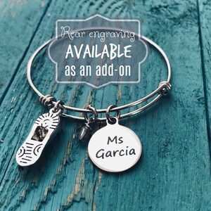 May include: Silver charm bracelet with a shoe charm, a crystal charm, and a round charm with the text "Ms Garcia". The bracelet is on a blue wooden surface. Rear engraving is available as an add-on.