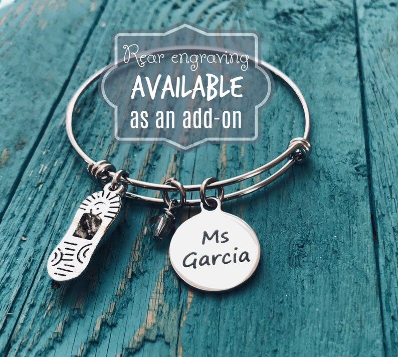 May include: Silver charm bracelet with a shoe charm, a crystal charm, and a round charm with the text "Ms Garcia". The bracelet is on a blue wooden surface. Rear engraving is available as an add-on.