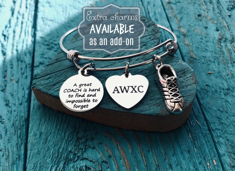 May include: A silver charm bracelet with three charms: a round charm with the text "A great COACH is hard to find and impossible to forget", a heart charm with the text "AWXC", and a shoe charm.