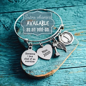 Pu&ograve; includere: Un braccialetto con ciondoli in argento con diversi ciondoli, tra cui un ciondolo a forma di cuore con il testo "Nov. 2020", un ciondolo rotondo con il testo "Someone I love died of an overdose", un ciondolo a forma di manubrio e un ciondolo a forma di ala. Il braccialetto &egrave; su una superficie di legno blu.