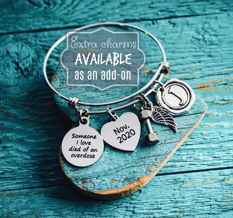 Pu&ograve; includere: Un braccialetto con ciondoli in argento con diversi ciondoli, tra cui un ciondolo a forma di cuore con il testo "Nov. 2020", un ciondolo rotondo con il testo "Someone I love died of an overdose", un ciondolo a forma di manubrio e un ciondolo a forma di ala. Il braccialetto &egrave; su una superficie di legno blu.