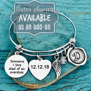 Pu&ograve; includere: Braccialetto in argento con un ciondolo a forma di cuore inciso con la data 12.12.18, un ciondolo rotondo con il testo "Someone I love died of an overdose", un ciondolo ad ala e un ciondolo con la lettera D. Il braccialetto &egrave; su una superficie di legno blu.