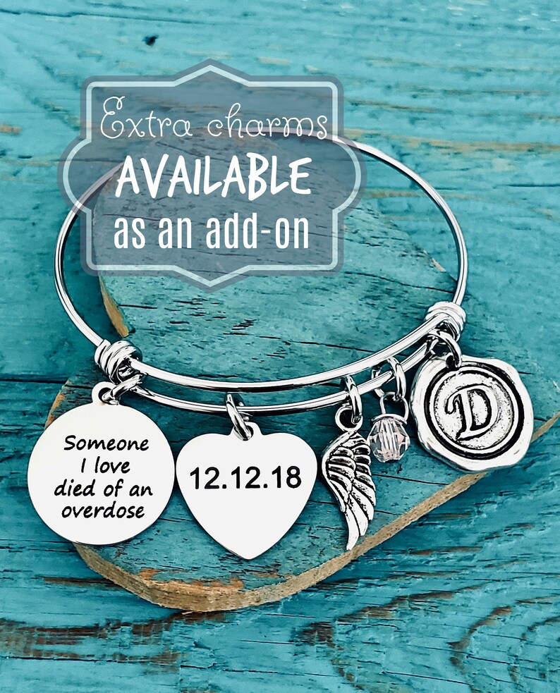 Pu&ograve; includere: Braccialetto in argento con un ciondolo a forma di cuore inciso con la data 12.12.18, un ciondolo rotondo con il testo "Someone I love died of an overdose", un ciondolo ad ala e un ciondolo con la lettera D. Il braccialetto &egrave; su una superficie di legno blu.